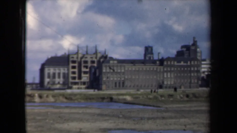 1946 Netherlands 8mm view of a scaffolded facade beside a long masonry building, with open muddy foreground and cloudy sky.