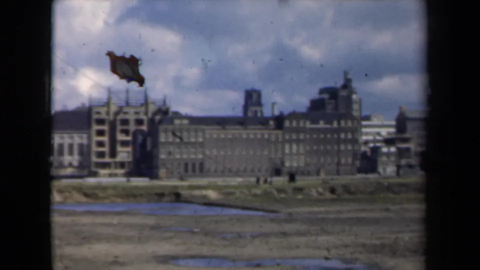1946 Netherlands 8mm frame showing scaffolded buildings and a distant tower over muddy ground, with a visible film damage mark near the sky.