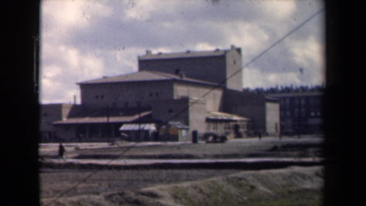 1946 Netherlands 8mm frame showing a blocky institutional building complex beyond a muddy open area, viewed through a window frame.