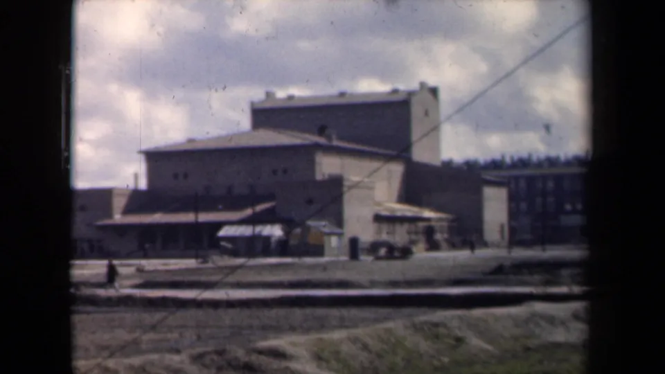 1946 Netherlands 8mm frame showing a blocky institutional building complex beyond a muddy open area, viewed through a window frame.