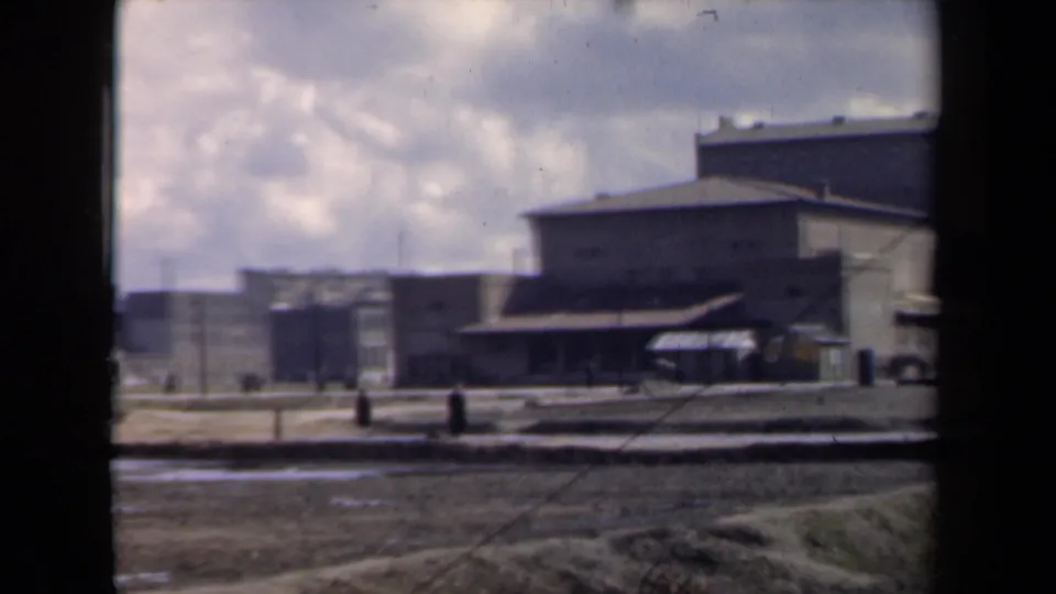 1946 Netherlands home-movie frame with distant pedestrians crossing a muddy courtyard before a large institutional building.