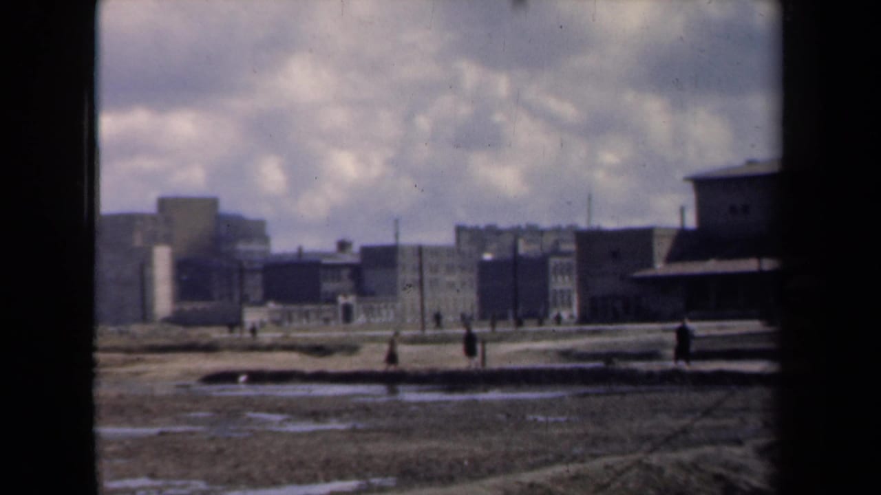 1946 Netherlands 8mm frame showing people crossing muddy open ground with factory buildings and smokestacks in the distance.