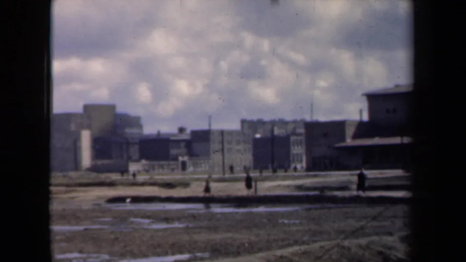 1946 Netherlands 8mm frame showing people crossing muddy open ground with factory buildings and smokestacks in the distance.