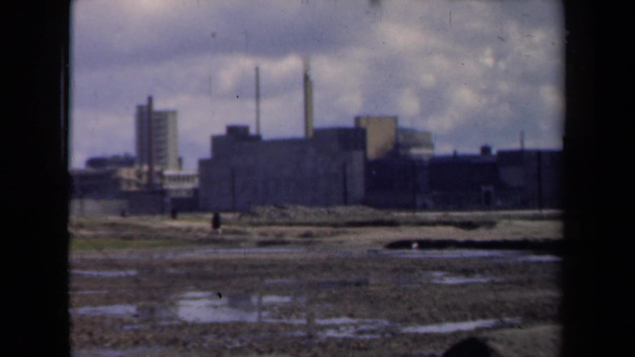 Factory buildings and smokestacks beyond wet open ground with small figures.