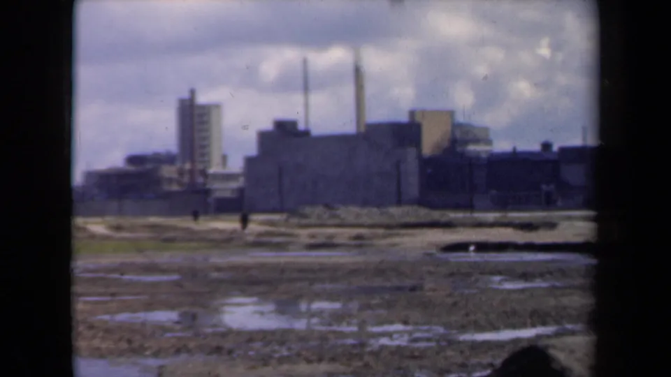 1940s Netherlands home-movie view of muddy ground, scattered walkers, and a large industrial complex beneath heavy clouds.