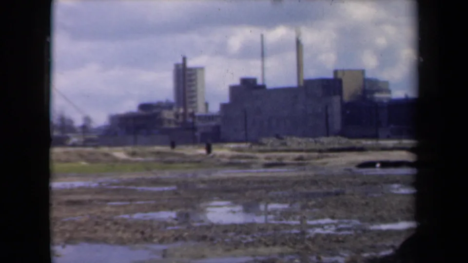Final 8mm frame of people moving through muddy open land in the Netherlands, set against distant factory buildings and smokestacks.