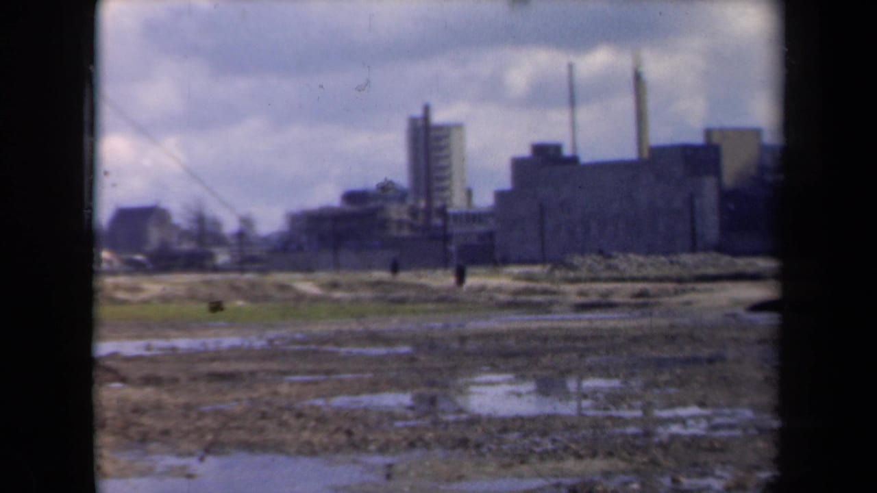 Distant factory buildings and smokestacks beyond a muddy, waterlogged field.
