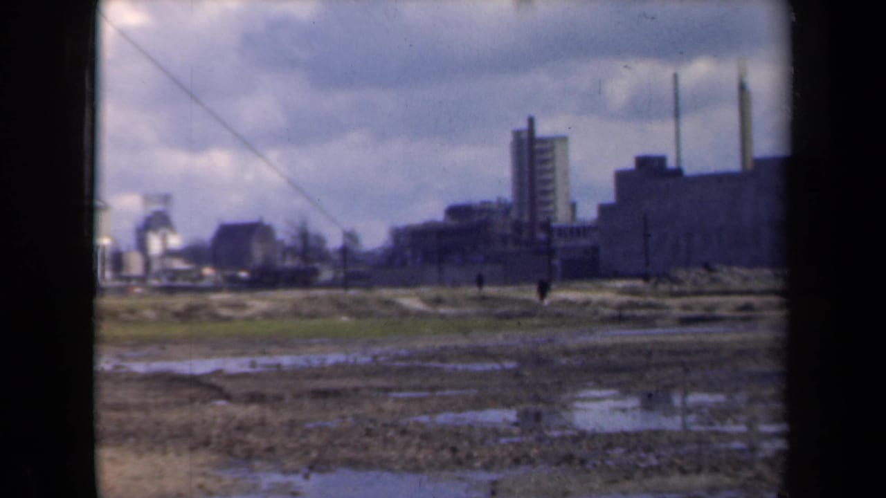 Industrial buildings and chimneys seen across wet ground with tiny figures in the distance.
