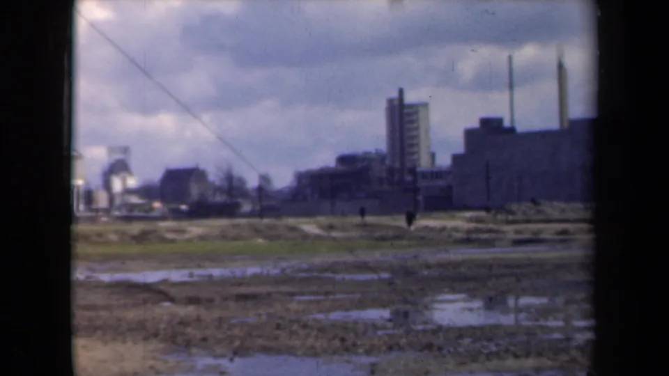 A distant industrial site in the Netherlands, filmed from across a muddy field with small pedestrians visible.