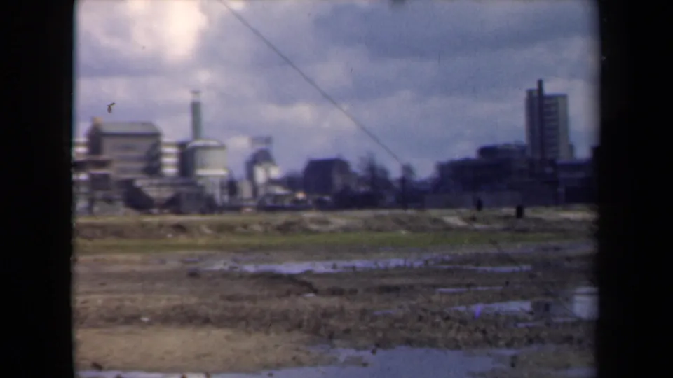 Distant 1946 Netherlands industrial complex viewed across muddy wetlands with one small figure visible.