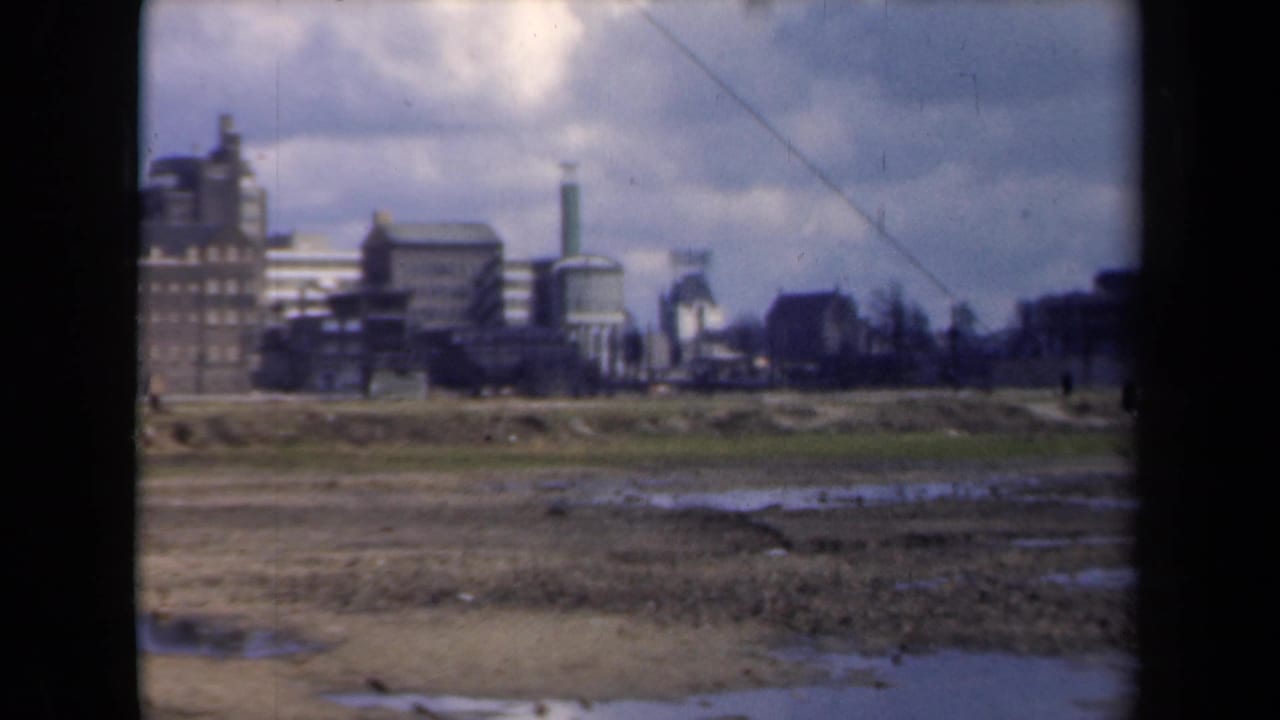 Industrial skyline beyond wet unused ground, with brick buildings, smokestacks, and cloudy sky.