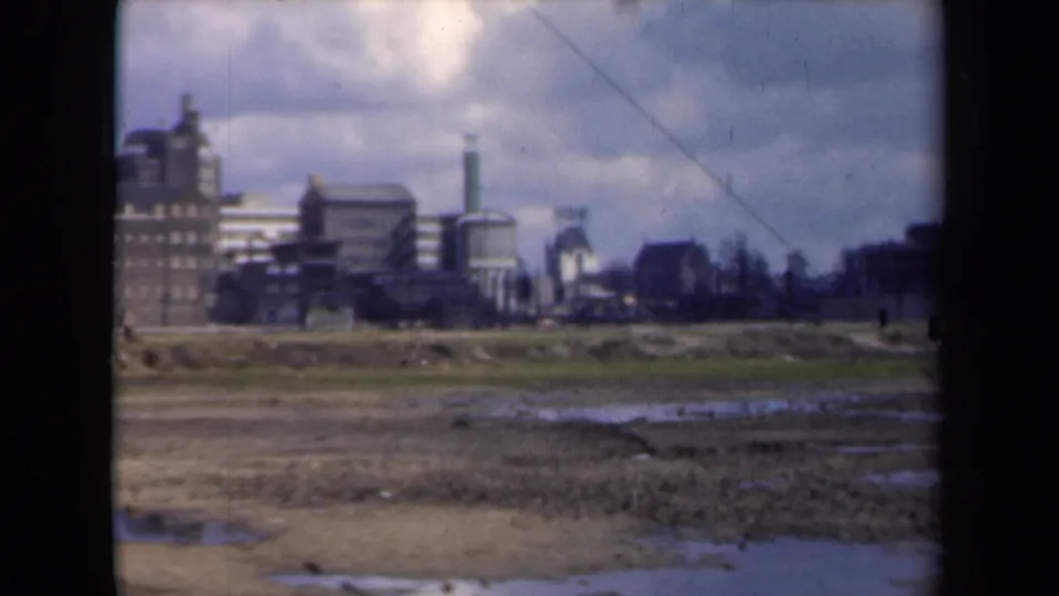 A 1946 Netherlands home-movie view looks over wet undeveloped land toward brick industrial buildings, stacks, and a tall tower.