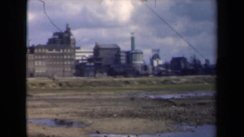 The 8mm frame shows marshy foreground terrain and a cluster of Dutch industrial buildings, with small distant people walking near the skyline.