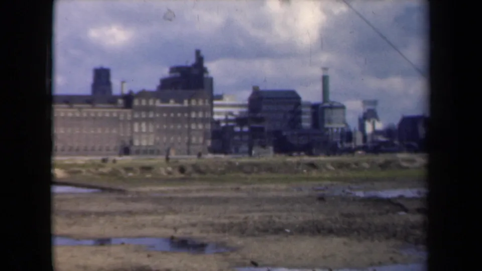 A 1946 Netherlands frame shows muddy land and puddles before a distant brick factory complex with smokestacks and small walking figures.
