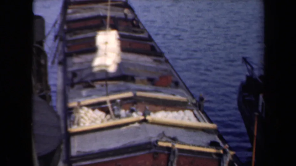 A yellow bundle descends toward the open deck of a docked cargo boat in the Netherlands archival harbor footage.