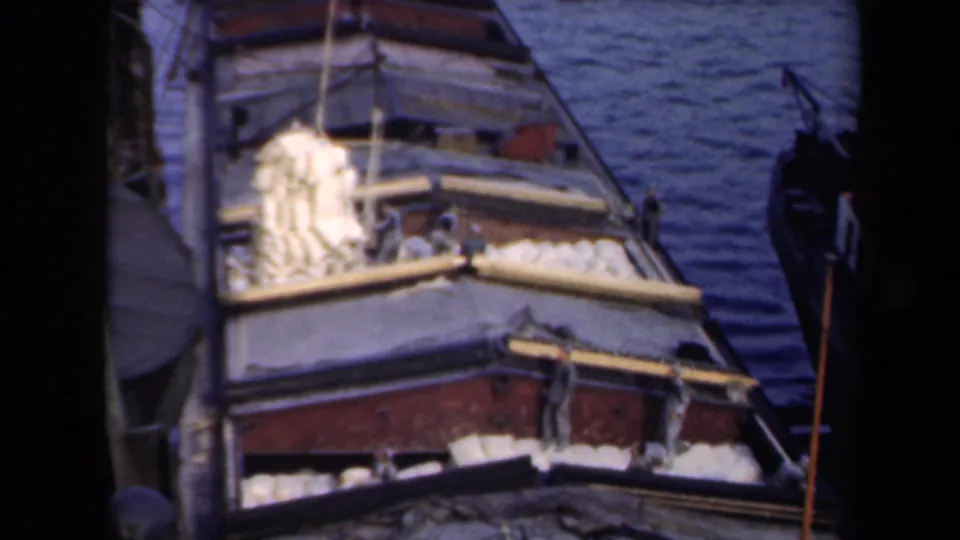 A docked cargo vessel carries yellow bundles on deck as another suspended bundle is lowered by rope, with small people visible onboard.