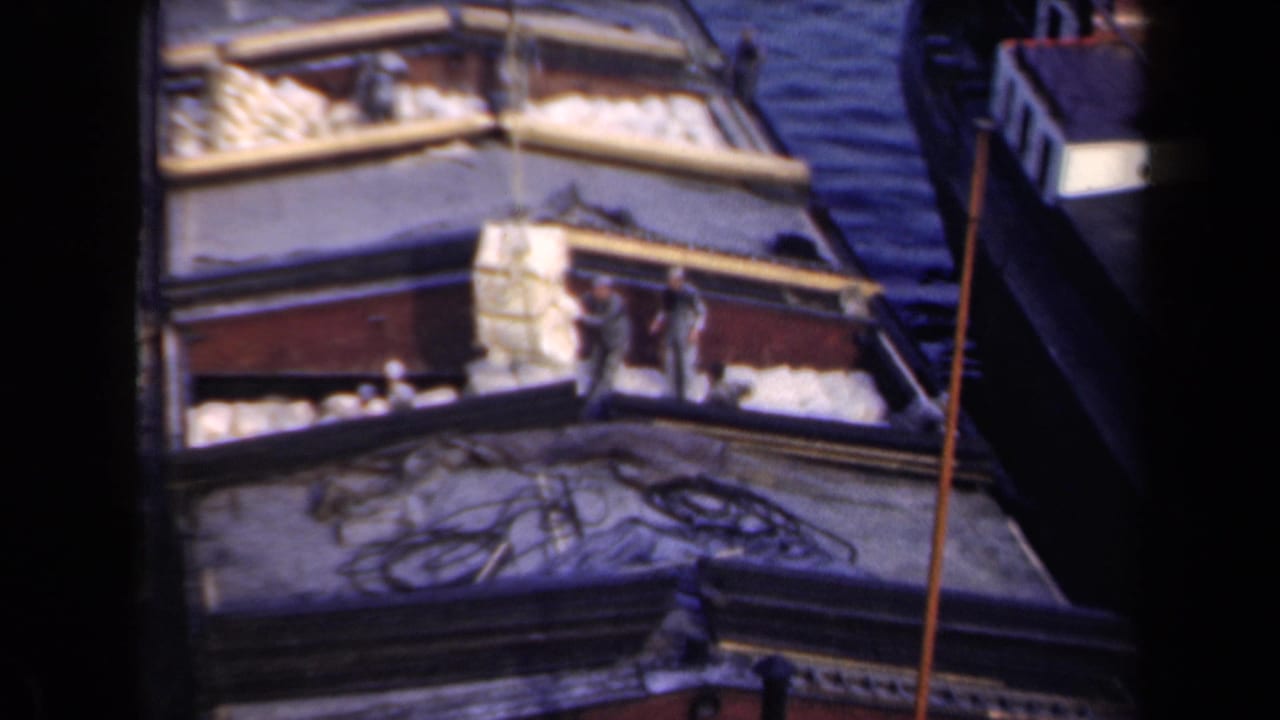 Workers stand on a barge deck with sacks, rope, and water visible around the boat.