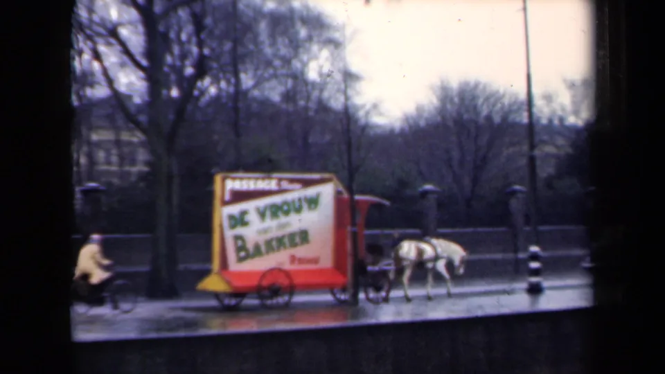 1946 Netherlands street scene with a cyclist passing a horse-drawn advertising cart beside wet pavement and bare trees.
