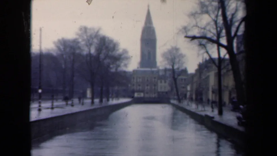 1946 Netherlands canal scene showing wet paths, bare trees, distant pedestrians, buildings, and a church spire at the far end.