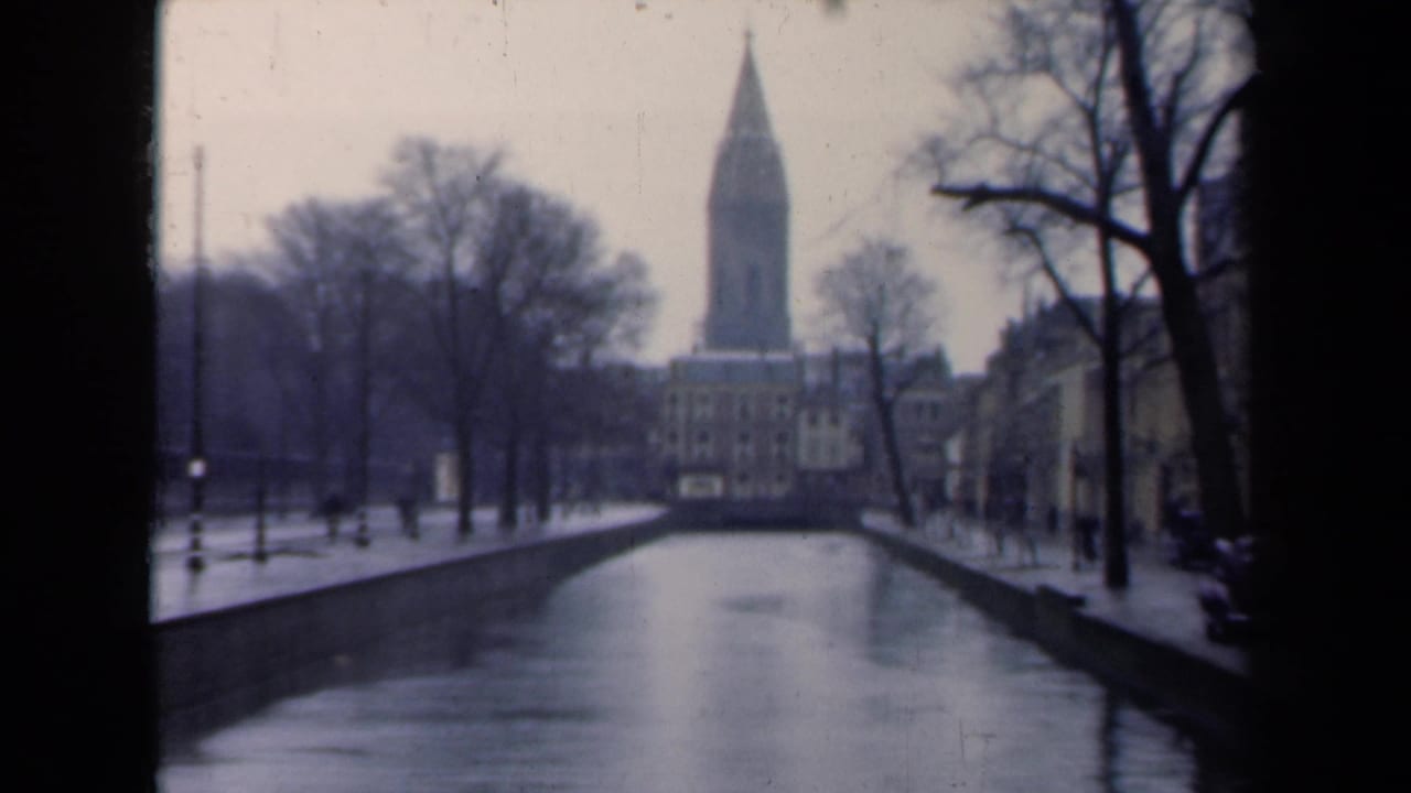 1946 Netherlands 8mm frame showing a canal, wet banks, leafless trees, buildings, and a tall spire in the distance.
