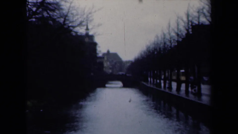 1946 Netherlands 8mm frame showing a canal, a small bridge, bare tree rows, and buildings partly silhouetted in soft light.