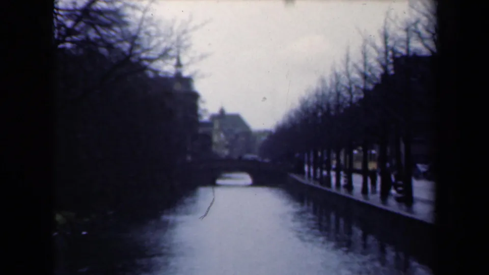 1946 Netherlands home-movie frame with a canal, arched bridge, bare trees, wet banks, and shadowed buildings beside the water.