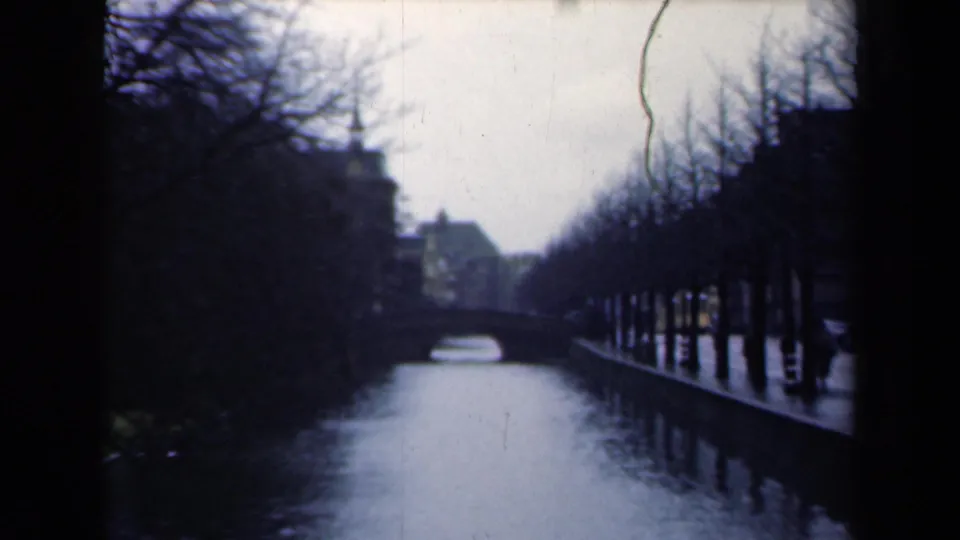 1946 Netherlands home-movie view of the canal near the Peace Palace, with bare trees, dark banks, buildings, and a small bridge ahead.