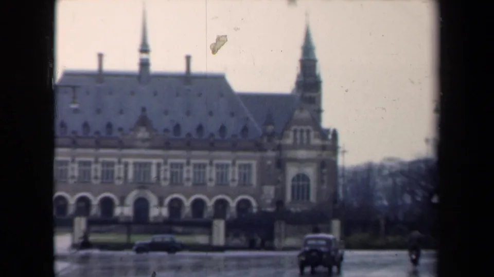 Wide 1946 Netherlands exterior of the Peace Palace on a rainy day, with wet pavement, parked cars, spires, arched windows, and distant pedestrians.