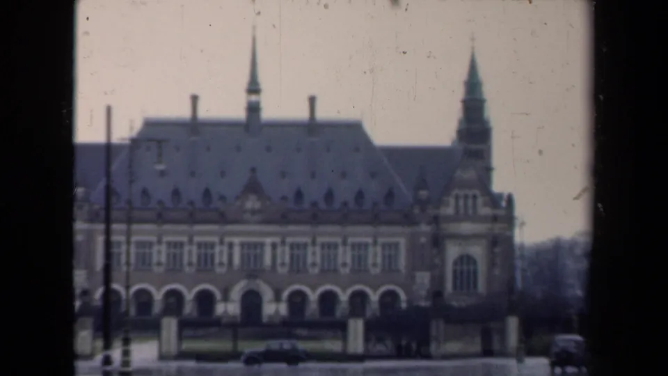 1946 Netherlands home-movie frame showing an ornate building behind a wet roadway, with mid-century cars passing.