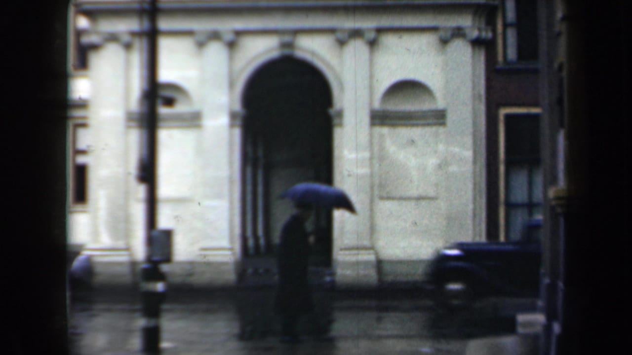 Rainy street outside a pale arched building with a person holding a dark umbrella.