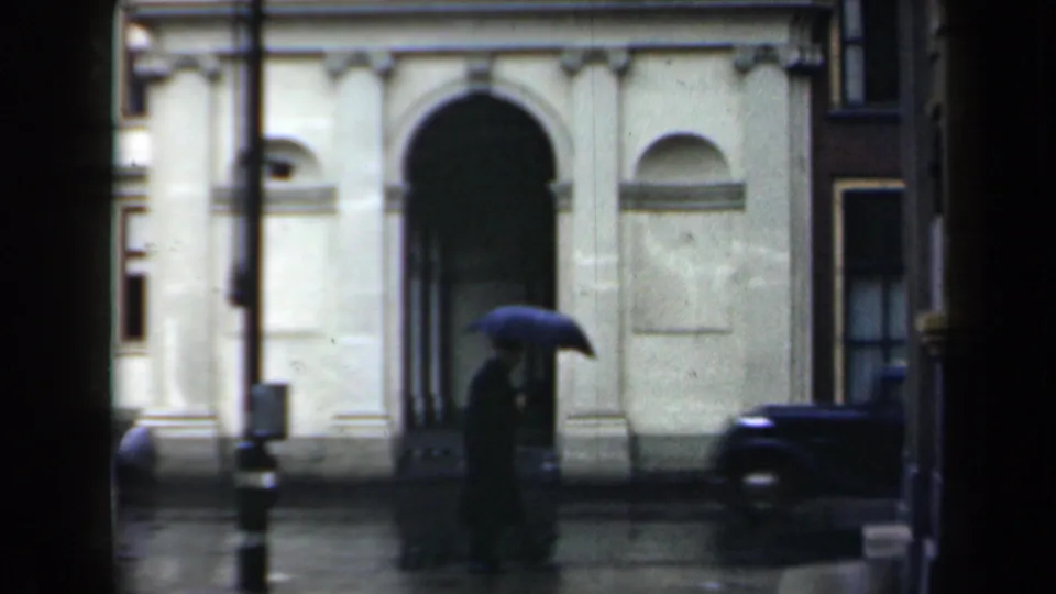 Rainy Netherlands street view of Queen Wilhelmina's palace facade, with an umbrella-bearing pedestrian and a dark car nearby.