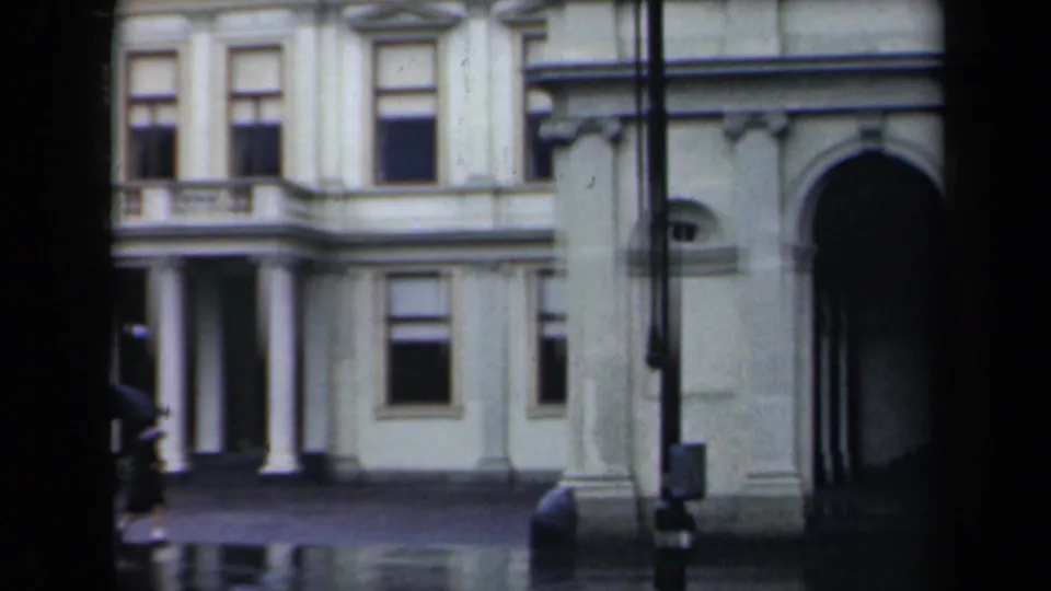 1946 Netherlands palace exterior showing balcony columns, tall windows, arched entrance, street furniture, and rain reflections.