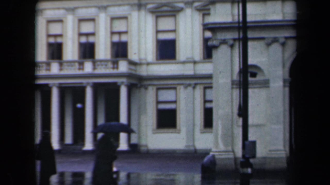 1946 Netherlands 8mm frame showing umbrella-carrying pedestrians before a white classical facade with columns and tall windows.