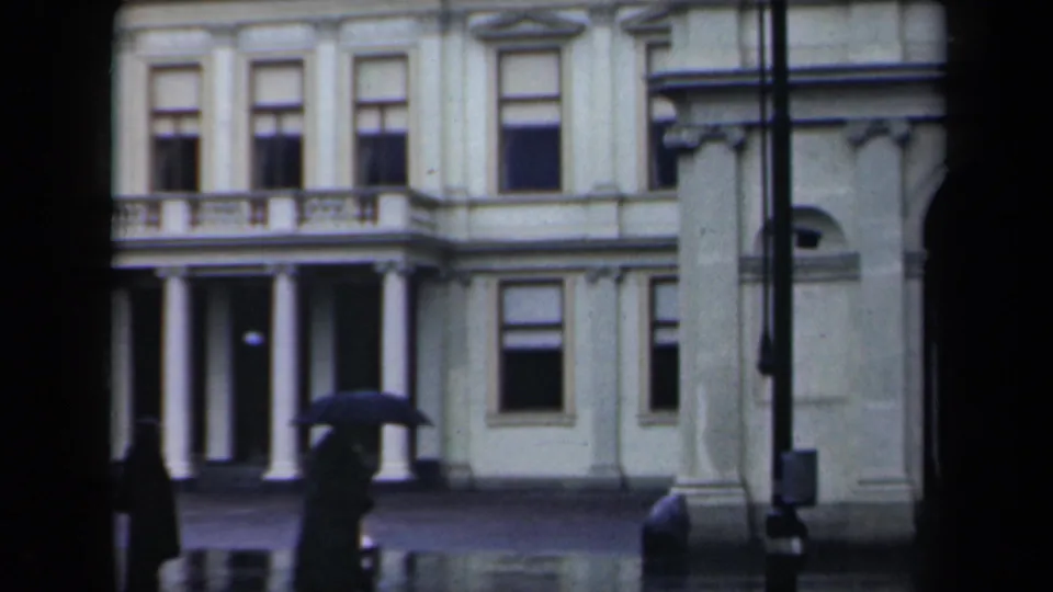 1946 Netherlands 8mm frame showing umbrella-carrying pedestrians before a white classical facade with columns and tall windows.