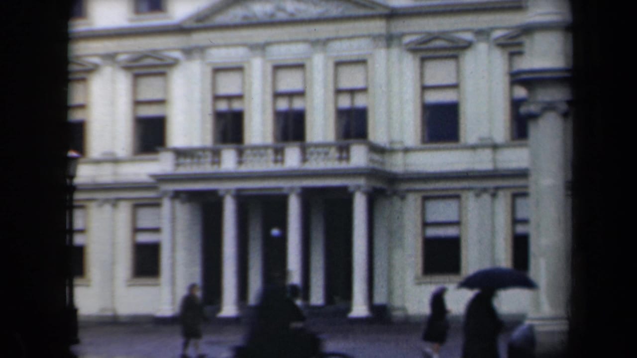 Rainy street view with people and umbrellas before a white building with columns.