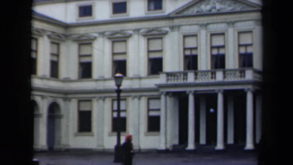 1946 Netherlands 8mm frame with a street lamp, a red head covering, pedestrians, and a white classical building in rain.