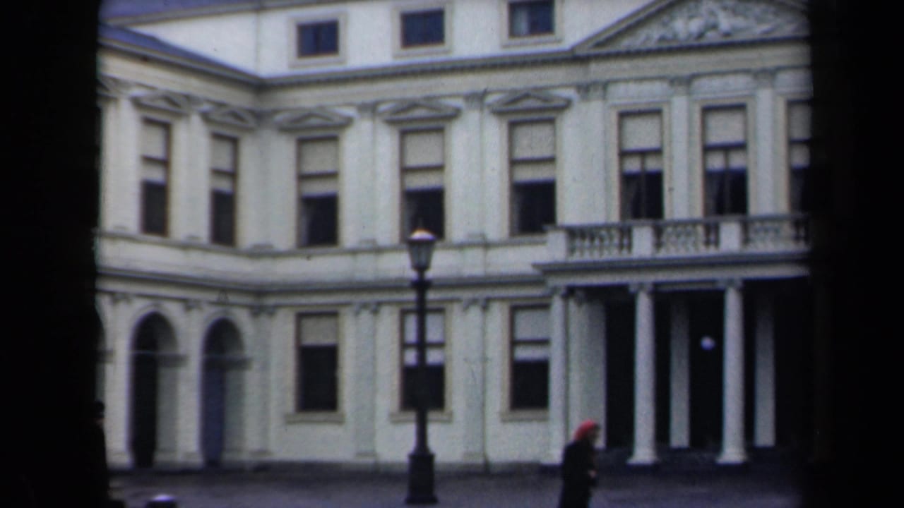 Classical building facade with columns, arched windows, streetlamp, and a pedestrian in a red hat.