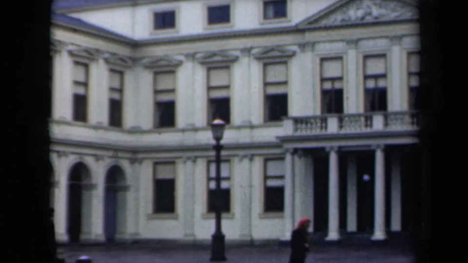 1946 Netherlands home-movie frame showing a pale classical building, columned entry, streetlamp, and a distant pedestrian in a red hat.