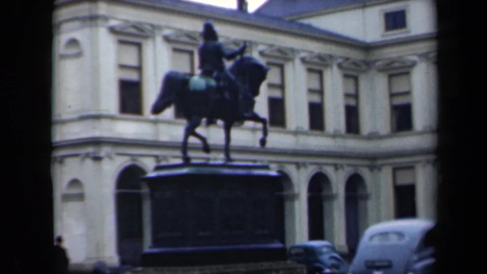 1946 Netherlands home-movie frame showing an equestrian statue, pale classical facade, arched windows, and period cars along the foreground.