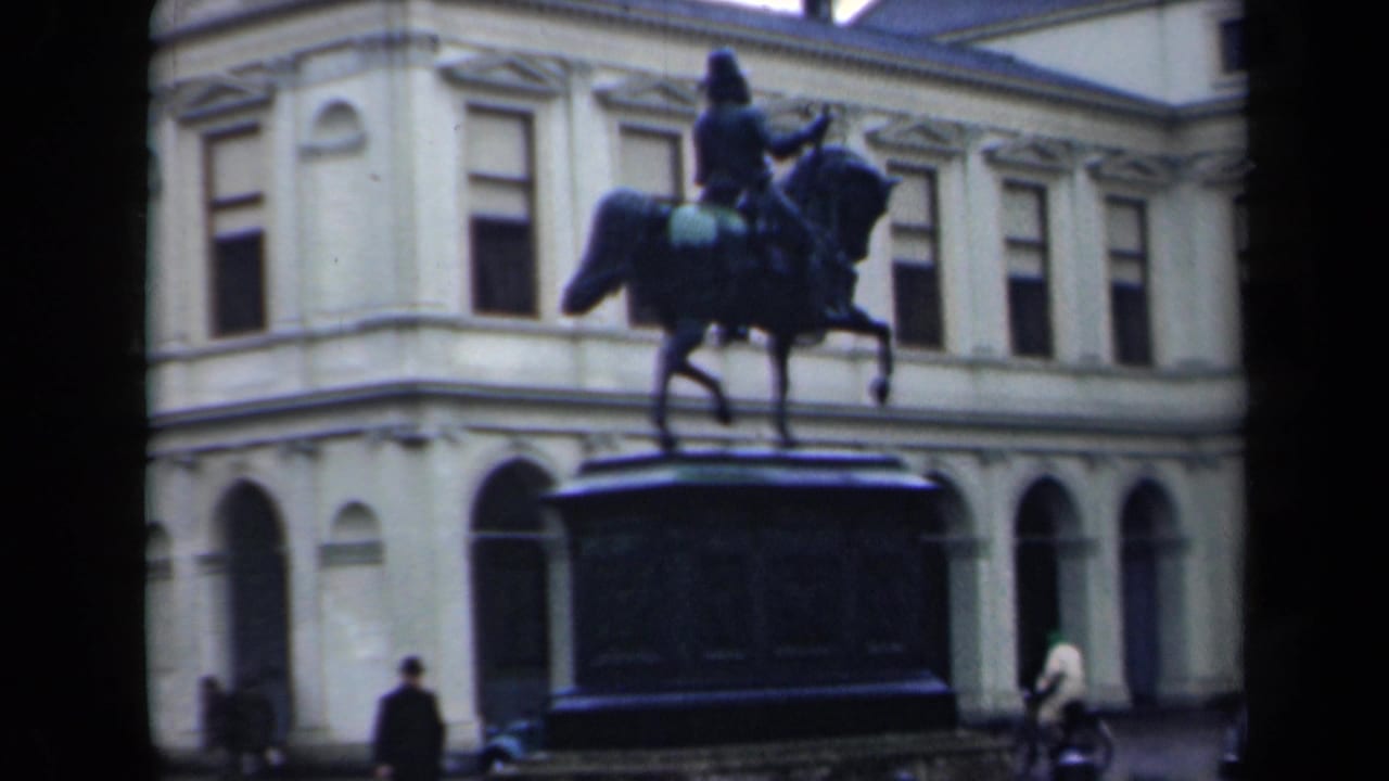 1946 Netherlands 8mm frame showing an equestrian statue in a public square before a pale classical building, with pedestrians at street level.