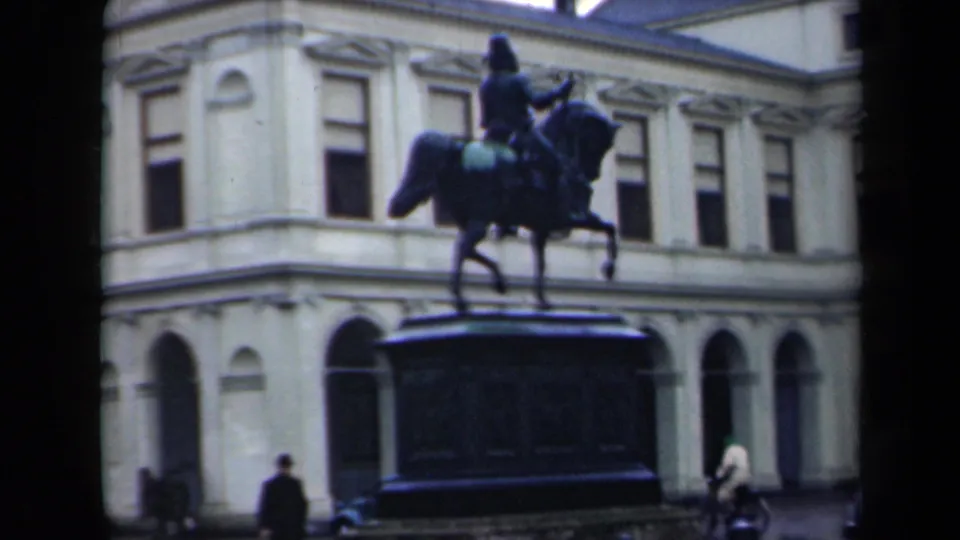 1946 Netherlands 8mm frame showing an equestrian statue in a public square before a pale classical building, with pedestrians at street level.