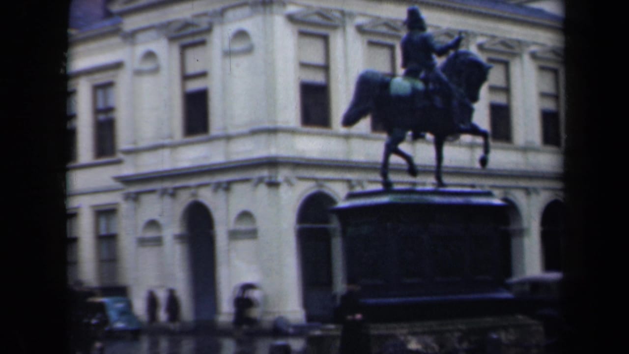 Equestrian statue centered in front of a pale building as people move through the square.
