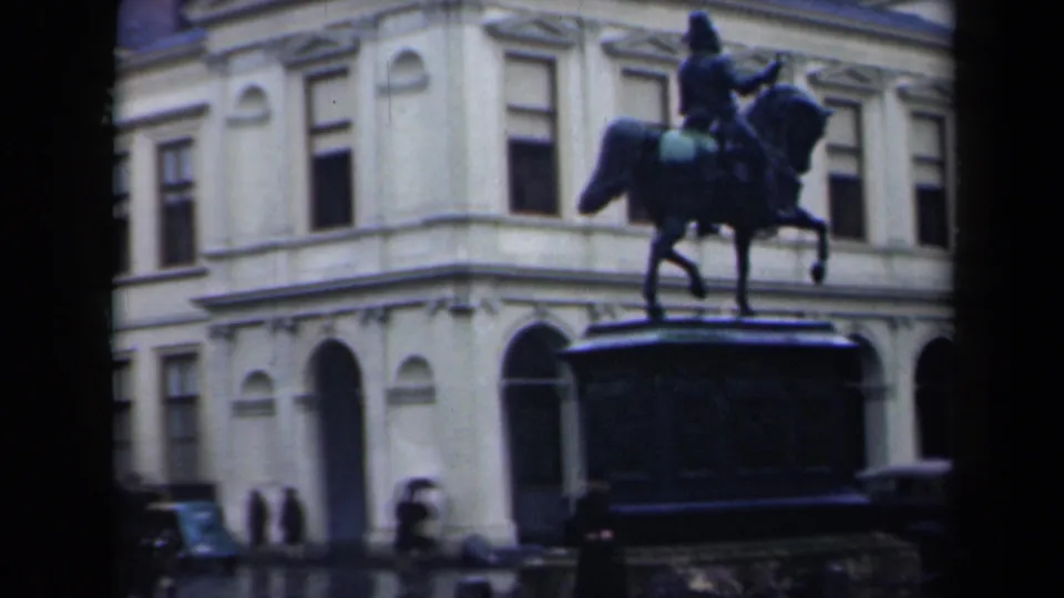 The statue and its large pedestal stand before a pale Dutch building, with small pedestrian figures and street details visible below.