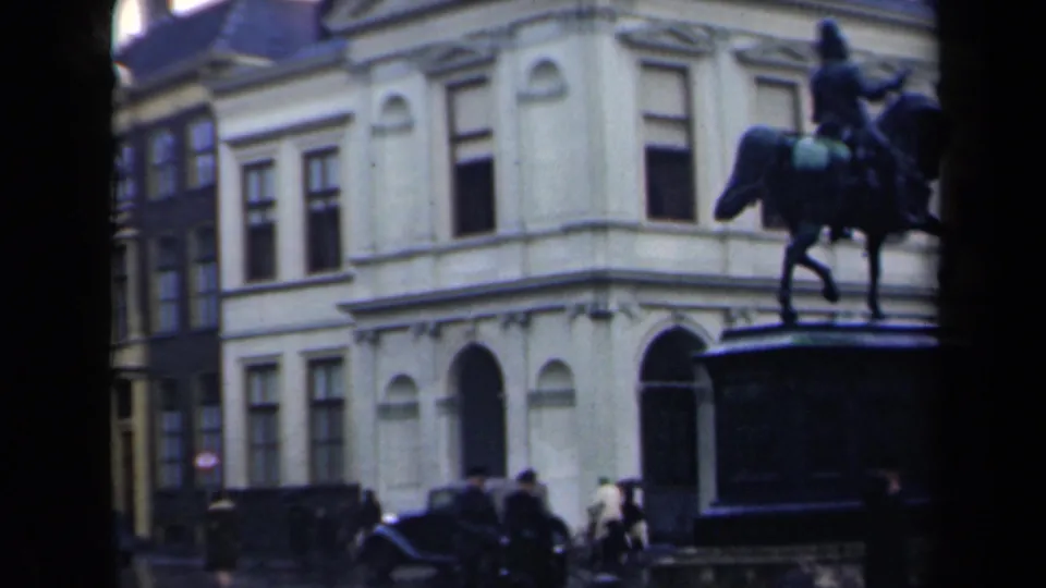 Handheld 8mm view of the equestrian monument from an angle, with period cars, pedestrians, and adjoining buildings in the square.
