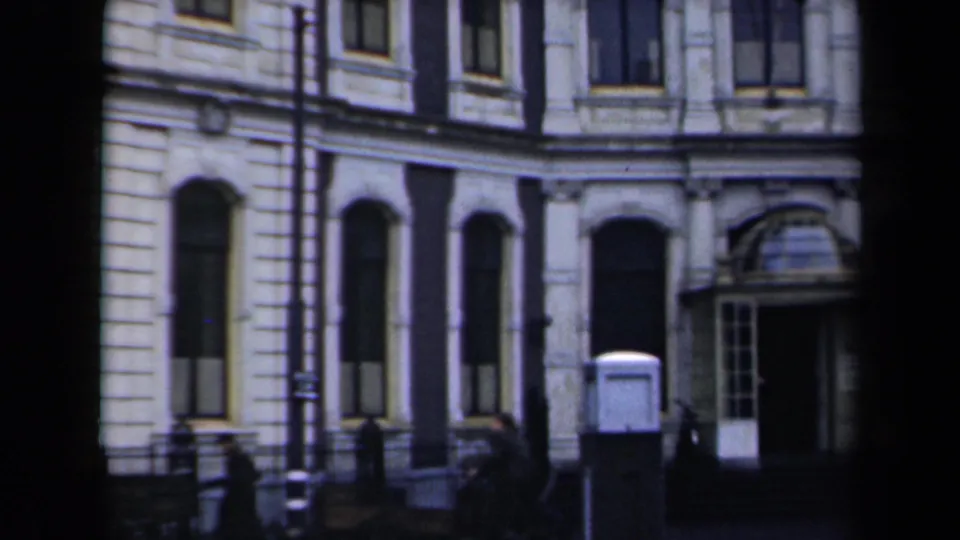 A later frame shifts from the statue to the nearby building facade, showing arched windows, a doorway, pedestrians, and street furniture.