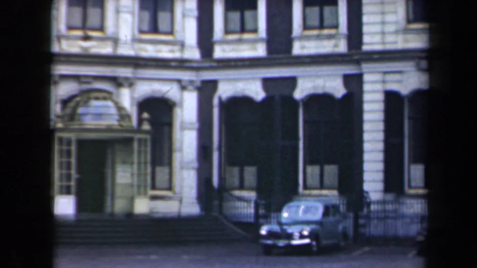 1946 Netherlands home-movie frame showing a grand stone facade, arched windows, entrance steps, iron railings, and a parked rounded sedan.