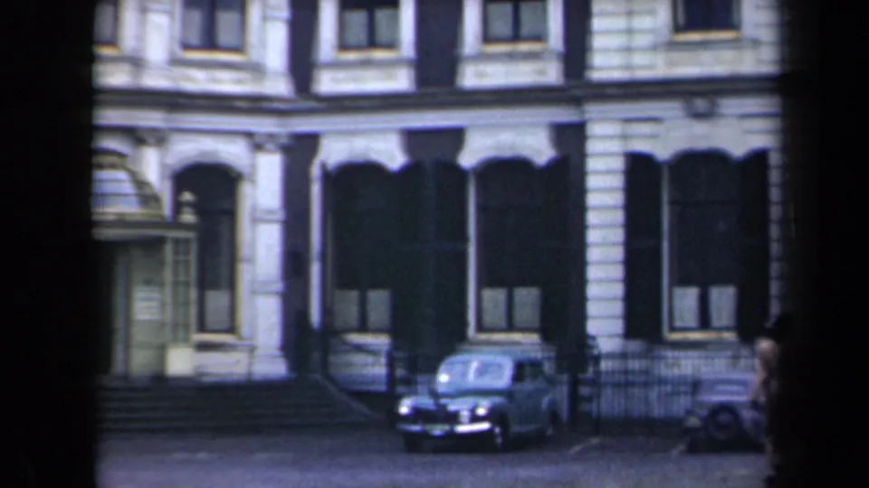 1940s Netherlands street view with an ornate masonry facade, dark awnings, railings, and a rounded parked car.