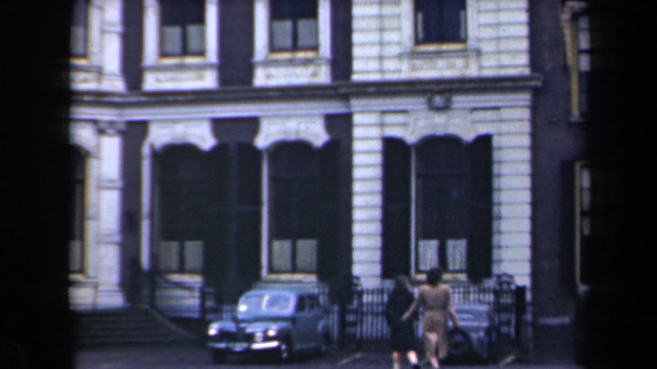 Two pedestrians pass a parked vintage car in front of an ornate building.