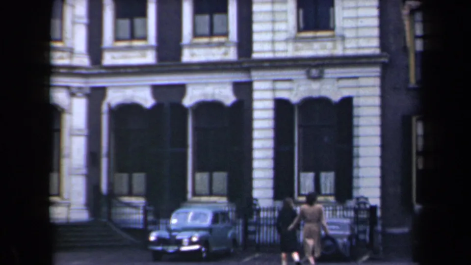 Two adults walk along a Netherlands street near a rounded parked car and a formal stone building facade.
