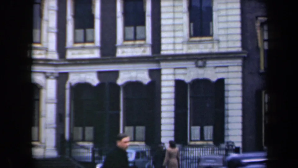 Street-level view of people, a bicycle, railings, and a parked car beside an ornate Netherlands building facade.